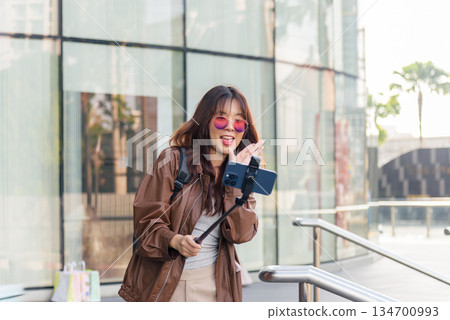 Asian woman enjoys shopping, people embrace fashion lifestyle with shopping bags, vlogging near modern mall, holding phone on stick, posing outside city building 134700993
