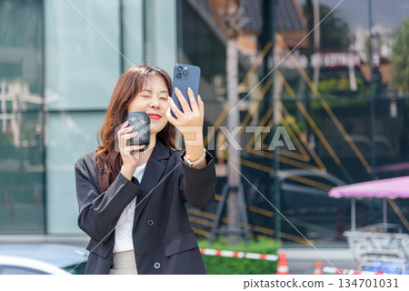 Young Asian woman, busy business work on street, smiles wearing suit, holds mobile phone taking self portrait, enjoys pleasant coffee moment, modern urban city background Young Asian woman, busy business work on street, smiles wearing suit, holds mobile phone taking self portrait, enjoys pleasant coffee moment, modern urban city background 134701031