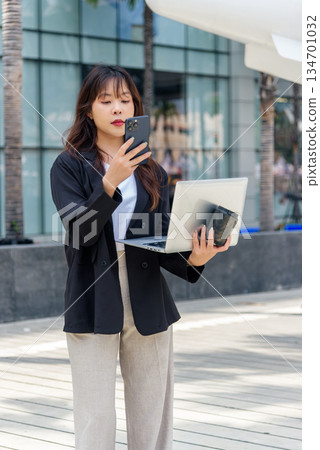 Young Asian business woman, busy professional, on city street works, wearing suit, managing digital tasks using smartphone, holding laptop, standing outdoor in urban environment communication Young Asian business woman, busy professional, on city street works, wearing suit, managing digital tasks using smartphone, holding laptop, standing outdoor in urban environment communication 134701032