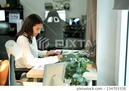 Female graphic designer working on logo creation with drawing tablet and color palette in modern bright office. 134703326