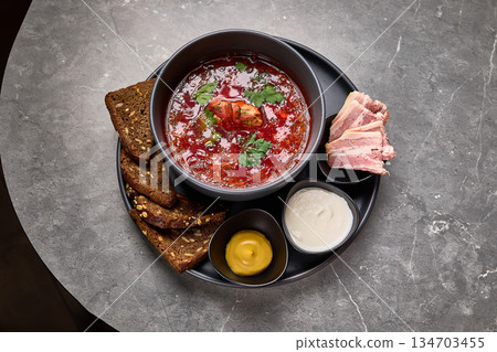 Traditional borscht with sour cream and rye bread on gray table, garnished with parsley and bacon slices, hearty meal concept Traditional borscht with sour cream and rye bread on gray table, garnished with parsley and bacon slices, hearty meal concept 134703455