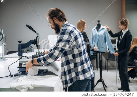 Guy in checkered shirt is working. People are by the sewing machines indoors 134704247