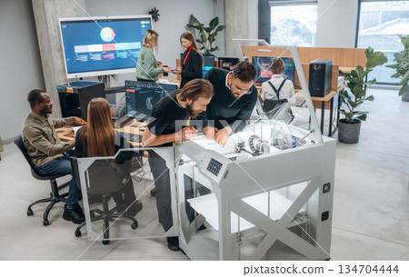 Group of people are standing around the 3d printer in office 134704444