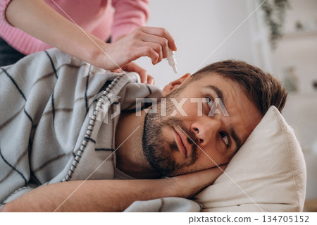 Funny facial expression. A man lies on a bed. A woman puts ear drops in his ear Funny facial expression. A man lies on a bed. A woman puts ear drops in his ear 134705152