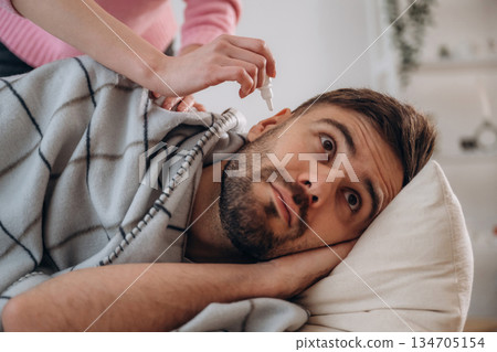 A man lies on a bed. A woman puts ear drops in his ear 134705154