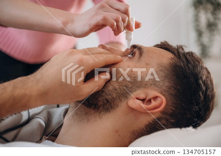 Man lies on a bed. A woman puts eyes drops in his eye Man lies on a bed. A woman puts eyes drops in his eye 134705157