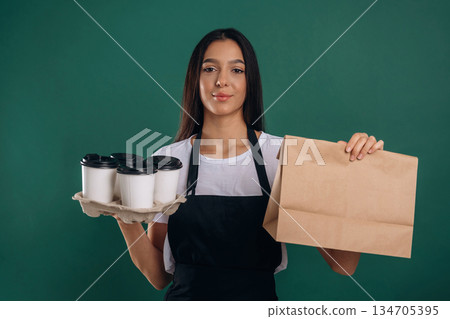 Cups of drinks and package in hands. Young beautiful barista is against background 134705395