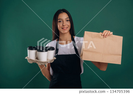 Cups of drinks and package in hands. Young beautiful barista is against background 134705396
