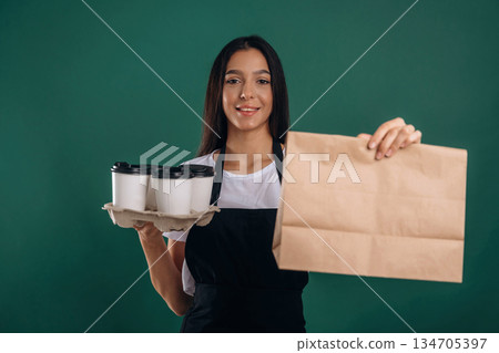 Cups of drinks and package in hands. Young beautiful barista is against background Cups of drinks and package in hands. Young beautiful barista is against background 134705397