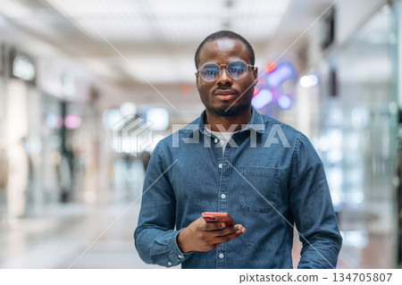 Front view, with smartphone in hand. Black man is in the hall of the supermarket 134705807
