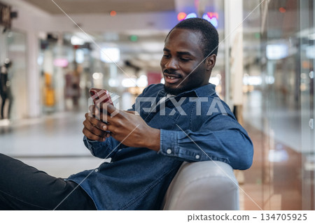 Sitting and holding smartphone. Black man is in the hall of the supermarket Sitting and holding smartphone. Black man is in the hall of the supermarket 134705925