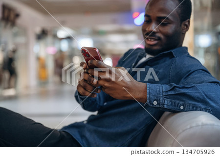 Sitting and holding smartphone. Black man is in the hall of the supermarket Sitting and holding smartphone. Black man is in the hall of the supermarket 134705926