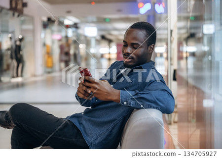 Sitting and holding smartphone. Black man is in the hall of the supermarket Sitting and holding smartphone. Black man is in the hall of the supermarket 134705927