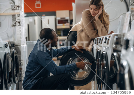 Sitting and opening. Man and woman are choosing washing machine in the store Sitting and opening. Man and woman are choosing washing machine in the store 134705999