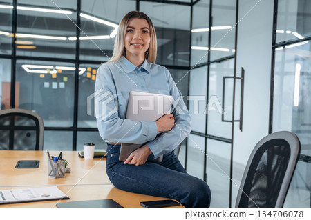 Posing, sitting on the table, holding laptop. Woman is in the modern office, formal wear Posing, sitting on the table, holding laptop. Woman is in the modern office, formal wear 134706078