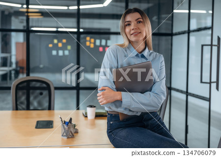 Posing, sitting on the table, holding laptop. Woman is in the modern office, formal wear Posing, sitting on the table, holding laptop. Woman is in the modern office, formal wear 134706079