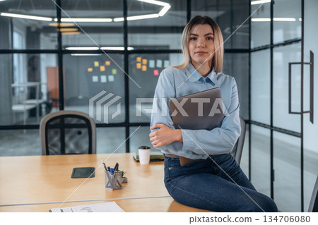 Posing, sitting on the table, holding laptop. Woman is in the modern office, formal wear Posing, sitting on the table, holding laptop. Woman is in the modern office, formal wear 134706080