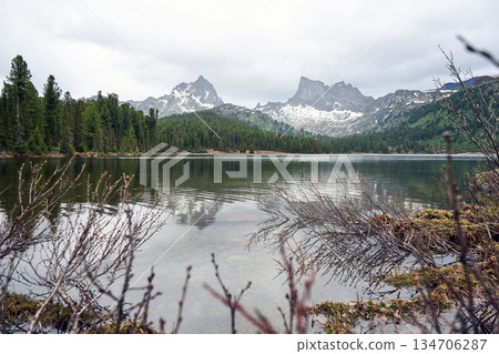 Calm mountain lake reflecting snow capped peaks, green forest under cloudy sky 134706287