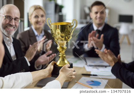 Business team celebrates success with golden trophy in office, applauding winner Business team celebrates success with golden trophy in office, applauding winner 134707816