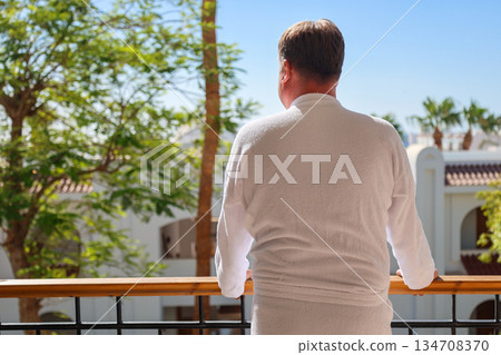 A man in a bathrobe stands on the terrace of a luxury house overlooking the sea and palm trees, rear view A man in a bathrobe stands on the terrace of a luxury house overlooking the sea and palm trees, rear view 134708370