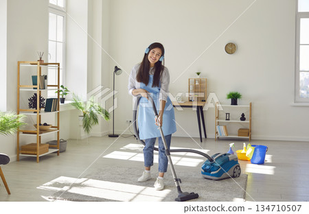 Happy young woman listening to music in headphones while vacuuming rug in living room 134710507