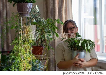 A woman stands in her home, expressing happiness and pleasure while tending to her plants. The woman is surrounded by a diverse collection of houseplants, reflecting a relaxed lifestyle. 134712282