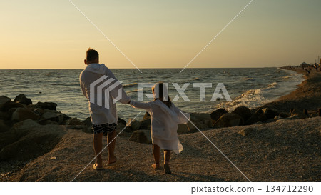 Two children, a boy and a girl, holding hands and walking together on a rocky beach towards the ocean during a warm sunset, symbolizing family connection, childhood, and summer freedom. Two children, a boy and a girl, holding hands and walking together on a rocky beach towards the ocean during a warm sunset, symbolizing family connection, childhood, and summer freedom. 134712290