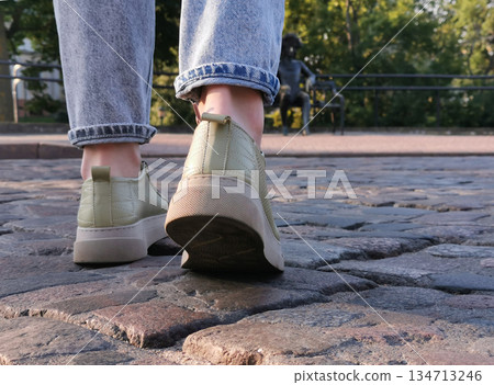 Close-up of someones feet in sneakers stepping on a cobblestone path, with a blurred statue in the background. 134713246