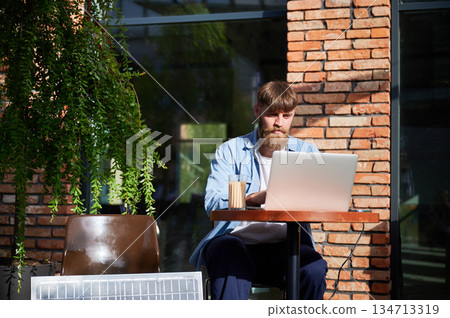 Man works on laptop at outdoor round table, with glass of iced coffee. Solar panel nearby, emphasizing sustainable, modern eco-friendly workspace that combines technology and sustainability. 134713319