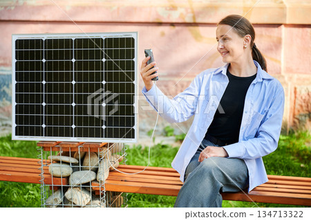Happy woman using smartphone connected to photovoltaic solar panel. Integration of sustainable renewable energy into everyday life, demonstrating practical use of solar power for charging devices. Happy woman using smartphone connected to photovoltaic solar panel. Integration of sustainable renewable energy into everyday life, demonstrating practical use of solar power for charging devices. 134713322