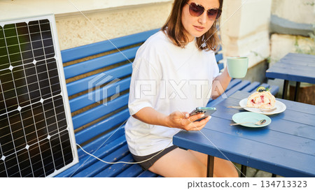 Girl drinking coffee while phone charging from solar panel. Cropped view of happy woman sitting next to portable solar battery charger. Integration of sustainable renewable energy into everyday life. 134713323