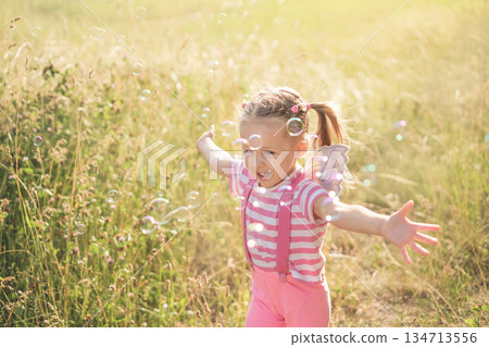A funny little girl with shiny fairy wings runs after soap bubbles flying in the wind in the rays of the setting sun. Happy childhood 134713556