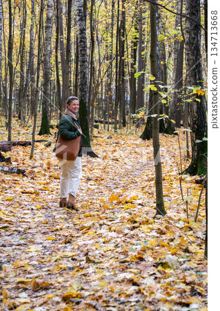 Smiling woman stands on a forest path covered with yellow autumn leaves, tall trees around, soft natural light, calm and peaceful mood 134713668