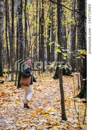 Smiling woman stands on a forest path covered with yellow autumn leaves, tall trees around, soft natural light, calm and peaceful mood 134713669
