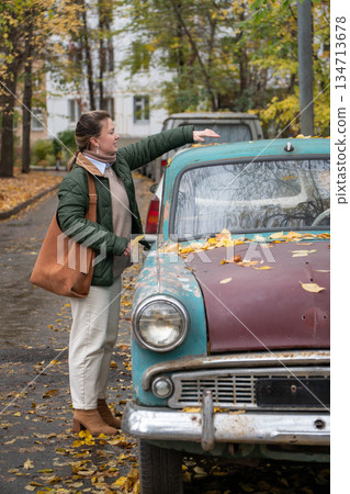 Woman smiles and gestures beside a vintage turquoise car covered with autumn leaves in residential courtyard, warm colors, playful mood Woman smiles and gestures beside a vintage turquoise car covered with autumn leaves in residential courtyard, warm colors, playful mood 134713678