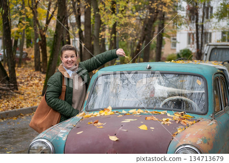 Woman smiles and gestures beside a vintage turquoise car covered with autumn leaves in residential courtyard, warm colors, playful mood Woman smiles and gestures beside a vintage turquoise car covered with autumn leaves in residential courtyard, warm colors, playful mood 134713679