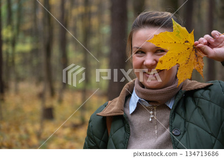 Woman holds a yellow maple leaf in front of her face in an autumn forest, wearing a green quilted jacket, beige sweater, and scarf 134713686