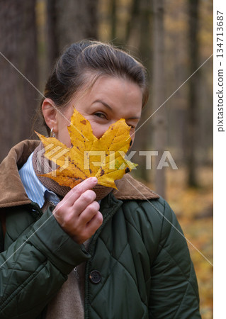 Woman holds a yellow maple leaf in front of her face in an autumn forest, wearing a green quilted jacket, beige sweater, and scarf Woman holds a yellow maple leaf in front of her face in an autumn forest, wearing a green quilted jacket, beige sweater, and scarf 134713687