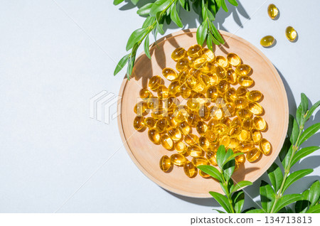 Yellow softgel vitamins capsules on a white wooden plate with green leaves on a blue background Yellow softgel vitamins capsules on a white wooden plate with green leaves on a blue background 134713813