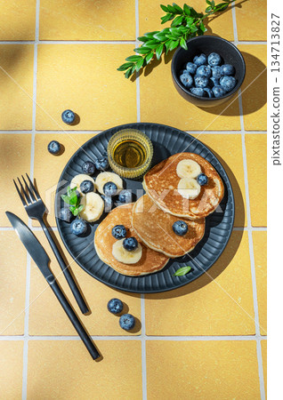 Pancakes with blueberry and banana on a plate on a yellow tile background with cutlery Pancakes with blueberry and banana on a plate on a yellow tile background with cutlery 134713827