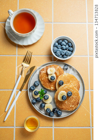 Pancakes with blueberry and banana on a plate on a yellow tile background with cup of tea Pancakes with blueberry and banana on a plate on a yellow tile background with cup of tea 134713828