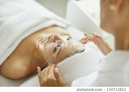 Female beautician applying moisturizing sheet mask on young woman face in beauty spa salon. Female beautician applying moisturizing sheet mask on young woman face in beauty spa salon. 134713852