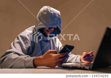 Hooded person engaging in cybercrime, performing online fraud and data theft using mobile phone, laptop and credit card in dark environment, representing internet security threats. Hooded person engaging in cybercrime, performing online fraud and data theft using mobile phone, laptop and credit card in dark environment, representing internet security threats. 134714255