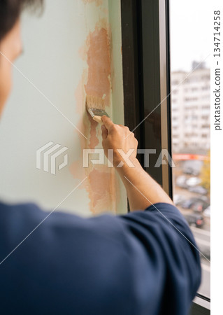Contractor male applying a sealant or primer coat to window frame wall with brush, preparing surface for painting or finishing during interior home improvement and repair project. Contractor male applying a sealant or primer coat to window frame wall with brush, preparing surface for painting or finishing during interior home improvement and repair project. 134714258