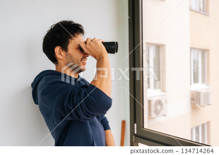 Curious young man looking through binoculars from bright apartment window, actively observing the adjacent building and city environment, engaging in surveillance or neighborhood watch. 134714264