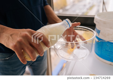 Close-up of male worker hands preparing white liquid primer by pouring from generic plastic bottle into empty mixing bowl, performing essential step for wall surface adhesion and renovation. Close-up of male worker hands preparing white liquid primer by pouring from generic plastic bottle into empty mixing bowl, performing essential step for wall surface adhesion and renovation. 134714269