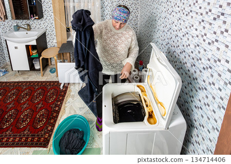 Freshly washed laundry is taken from drum of automatic top loading washing machine and moved into plastic basin, woman about 50 years old does housework. Freshly washed laundry is taken from drum of automatic top loading washing machine and moved into plastic basin, woman about 50 years old does housework. 134714406