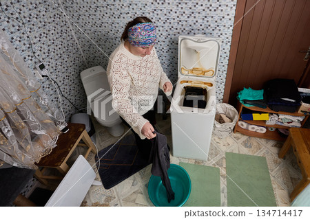 Woman takes washed laundry out of drum of vertical washing machine and puts it into plastic basin. Woman takes washed laundry out of drum of vertical washing machine and puts it into plastic basin. 134714417