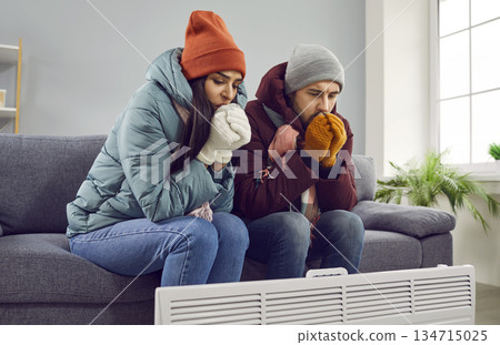 Young attractive couple are sitting on the sofa at home in warm clothes and warming their hands with heater in the living room, freezing from the cold. Heating problems, room is cold, people freezing. 134715025