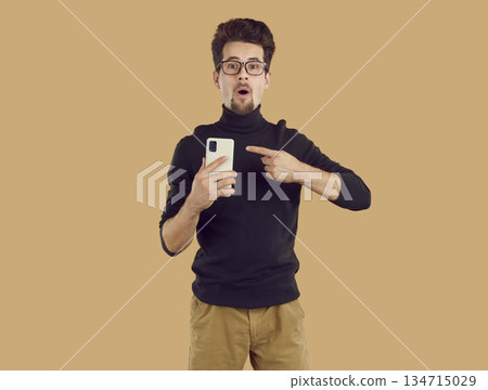 Amazed young brunet man in black turtleneck is pointing at smartphone on beige background. 134715029